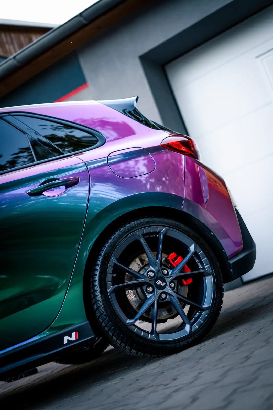 Rear wing and wheel in the same colour-shift (chameleon) film outdoors by a garage, an N logo legible on the caliper and side trim.