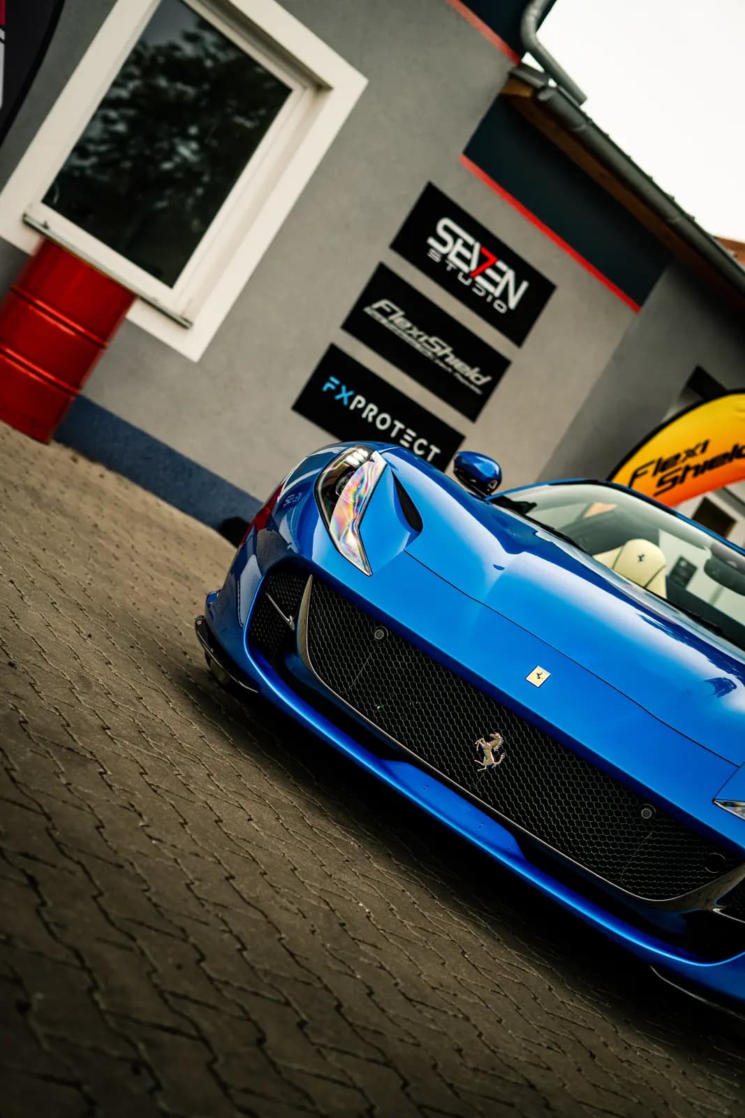Blue Ferrari with the prancing horse on the grille, parked on outdoor paving in front of a building with Seven Studio, FlexiShield and FXPROTECT signs.