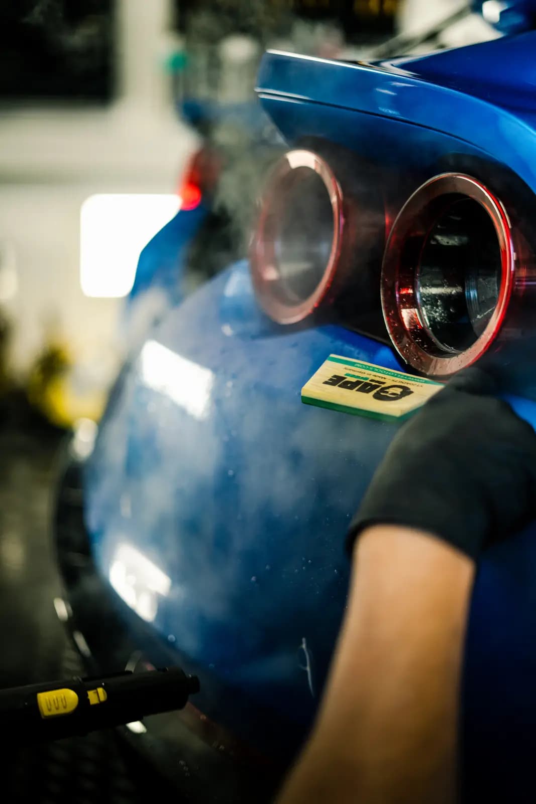 Rear of a blue car with round lights in red surrounds; a technician in a black glove applying yellow-green tape marked TAPE to the body near the light.