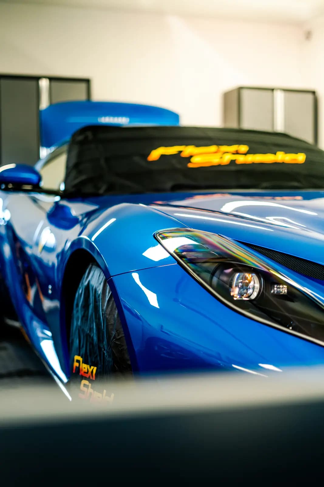 Front three-quarter view of a blue car with a windscreen cover printed “Flexi Shield” and “PPF Shield”, grey cabinets and another blue car blurred behind.
