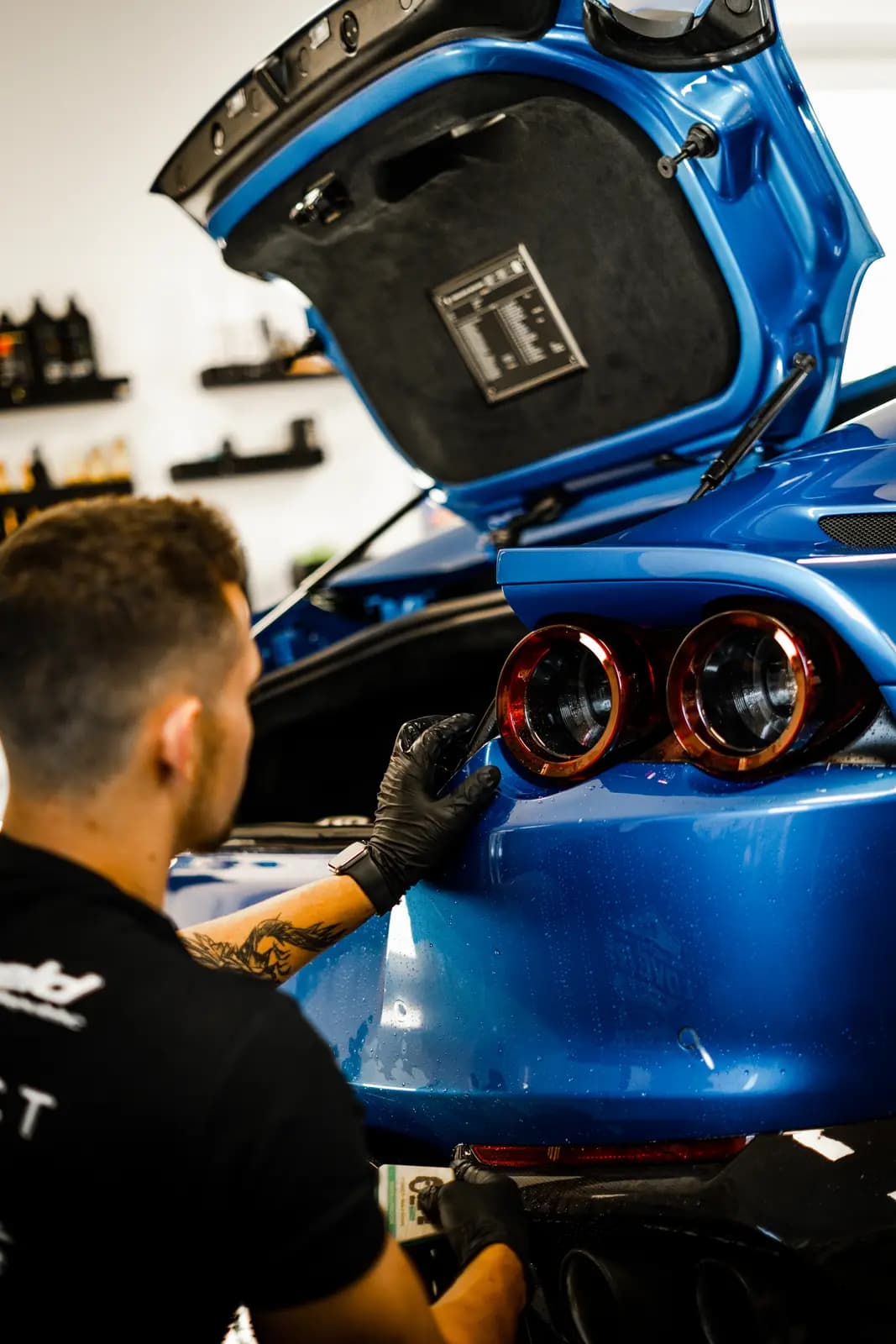 Man in a black shirt and black gloves leaning over the open boot of a blue car, a hand on the rear bumper beside the round lights in red surrounds; a shelf of detailing bottles behind.