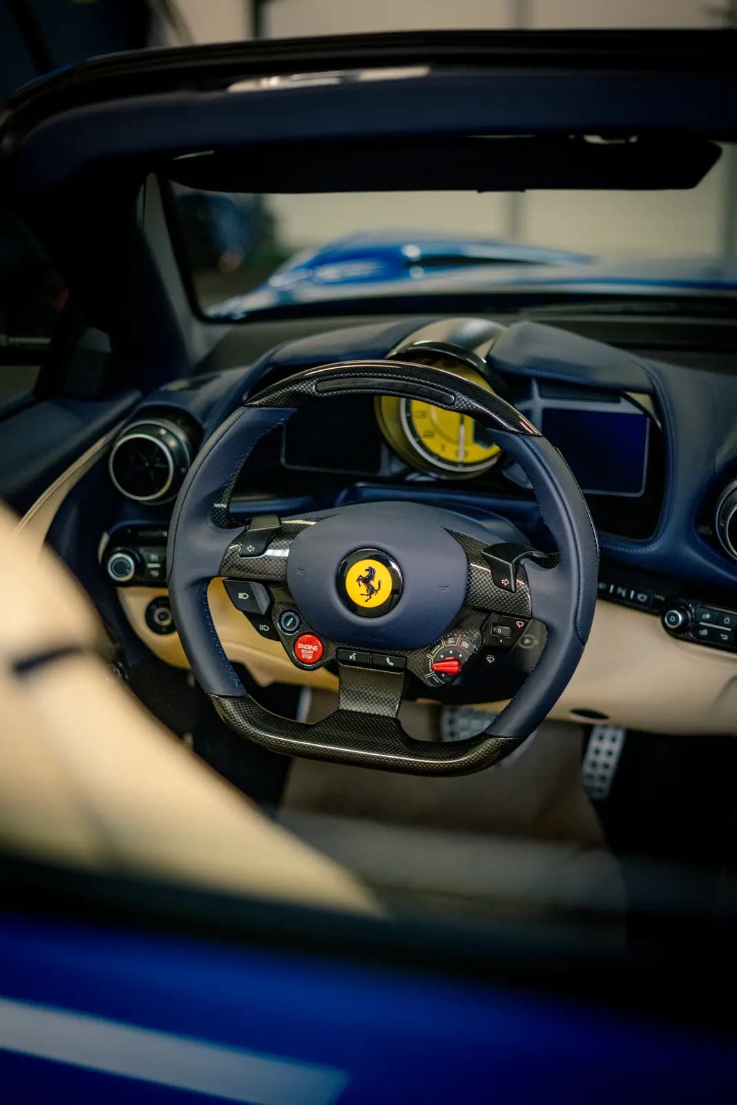 Steering wheel with the yellow Ferrari badge and carbon trim, a red ENGINE START button below and a yellow dial on the instrument panel; another blue car blurred behind.