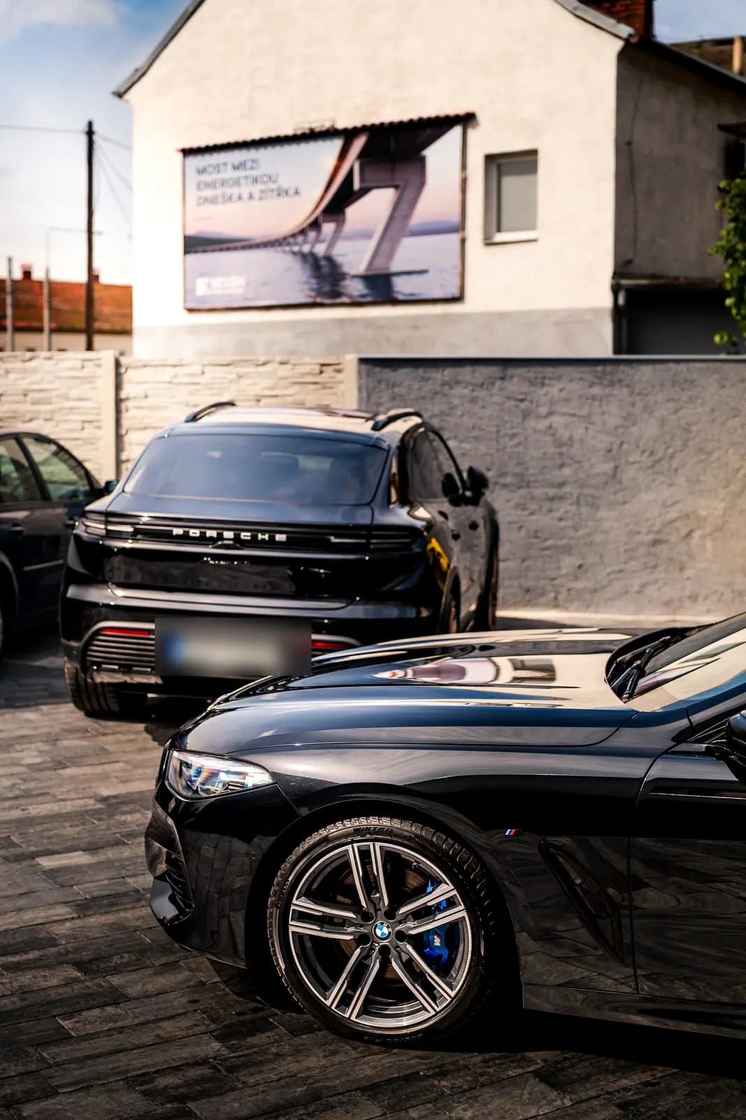 Rear of a black Porsche Cayenne with Porsche lettering, the nose of another black car in the foreground, outdoors in front of a building with a billboard.