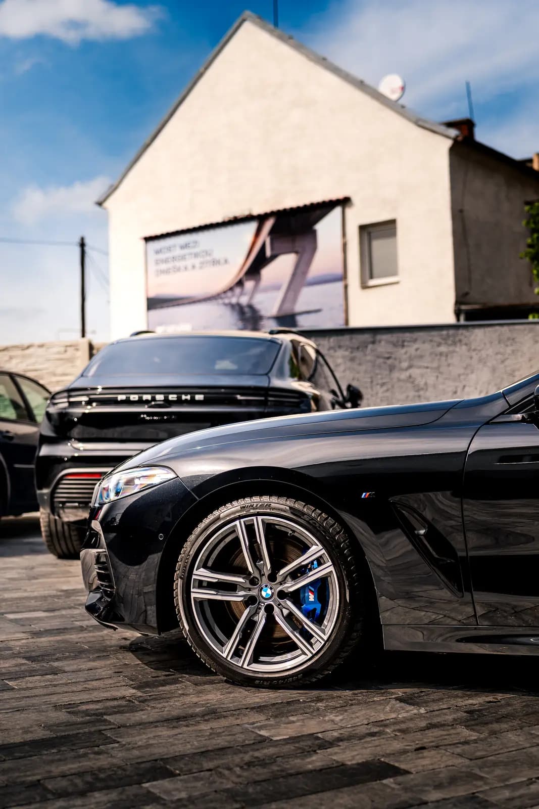 Front wing of a black BMW with the M badge and a blue brake caliper, a black Porsche Cayenne behind, on outdoor paving.