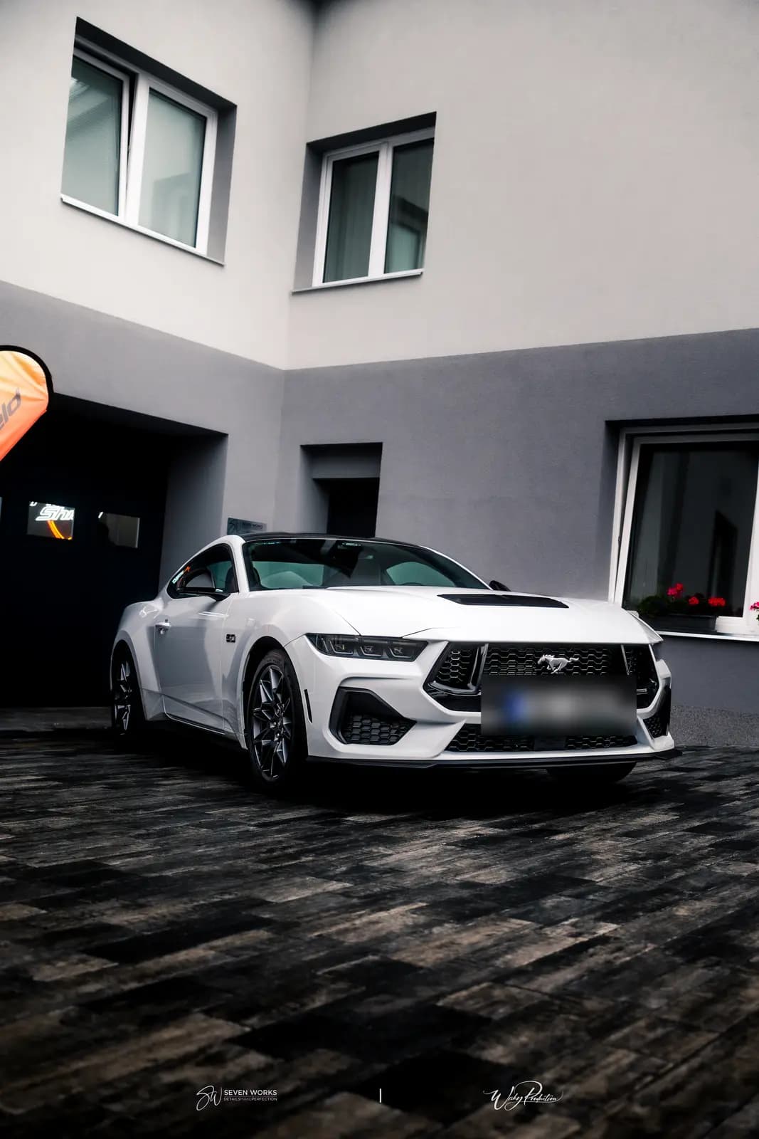 White sports coupe parked outdoors in a paved yard in front of a building with garage doors, an orange flag at left.