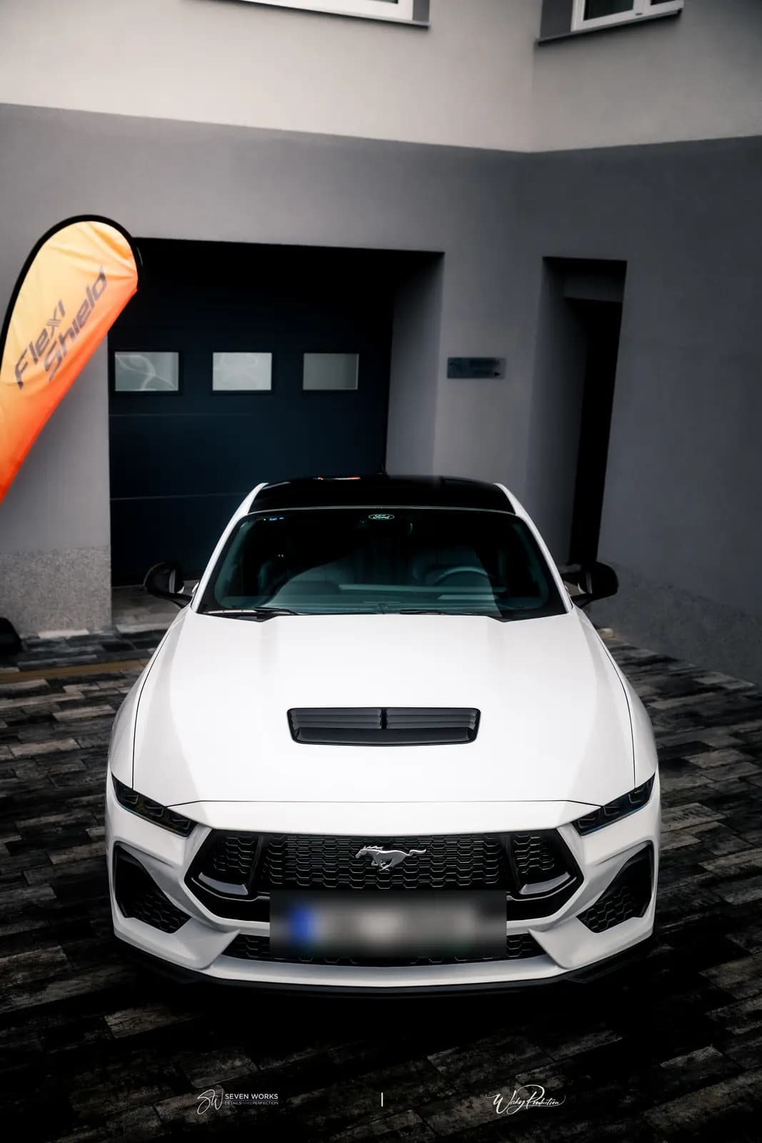 Head-on view of a white sports coupe with a black roof and the pony badge on the grille, an orange Flexi Shield flag beside it.