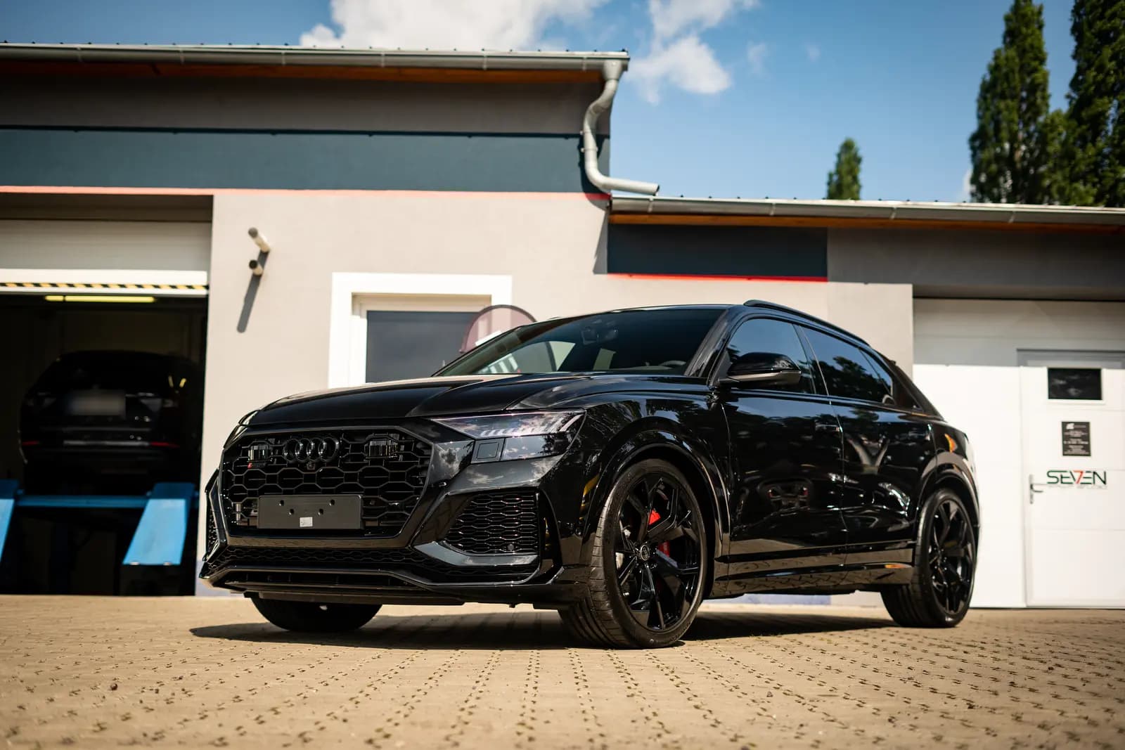 Black Audi SUV from the front and side with the Audi badge on the grille, parked on paving in front of a building with Seven signage and an open garage.
