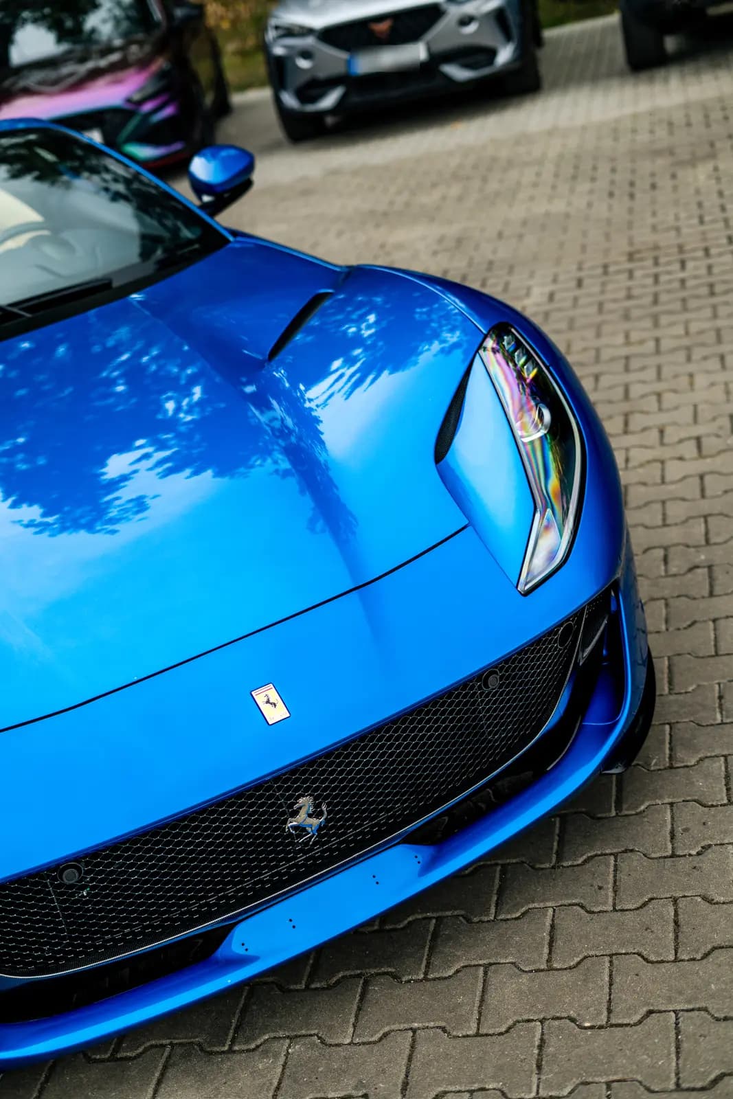 Detail přední kapoty a světlometu modrého Ferrari se znakem koně na masce, v pozadí rozmazaná auta včetně barvoměnného vozu.