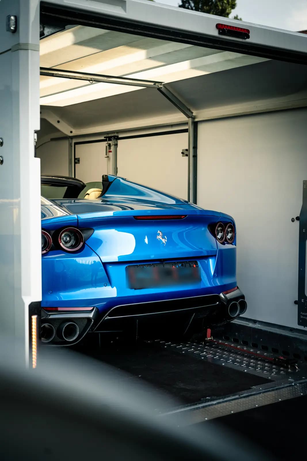 Zadní pohled na modrý vůz s emblémem Ferrari na víku kufru a čtyřmi koncovkami výfuku, stojící na nájezdové rampě uzavřeného přepravního návěsu.