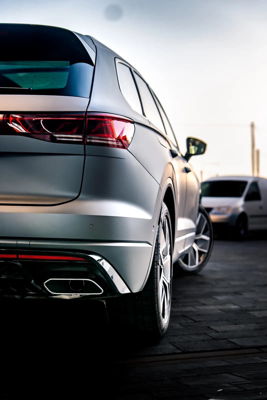 Volkswagen Touareg v matné šedé fólii zezadu a ze strany, se zadním světlem a koncovkou výfuku; v pozadí rozostřená dodávka.