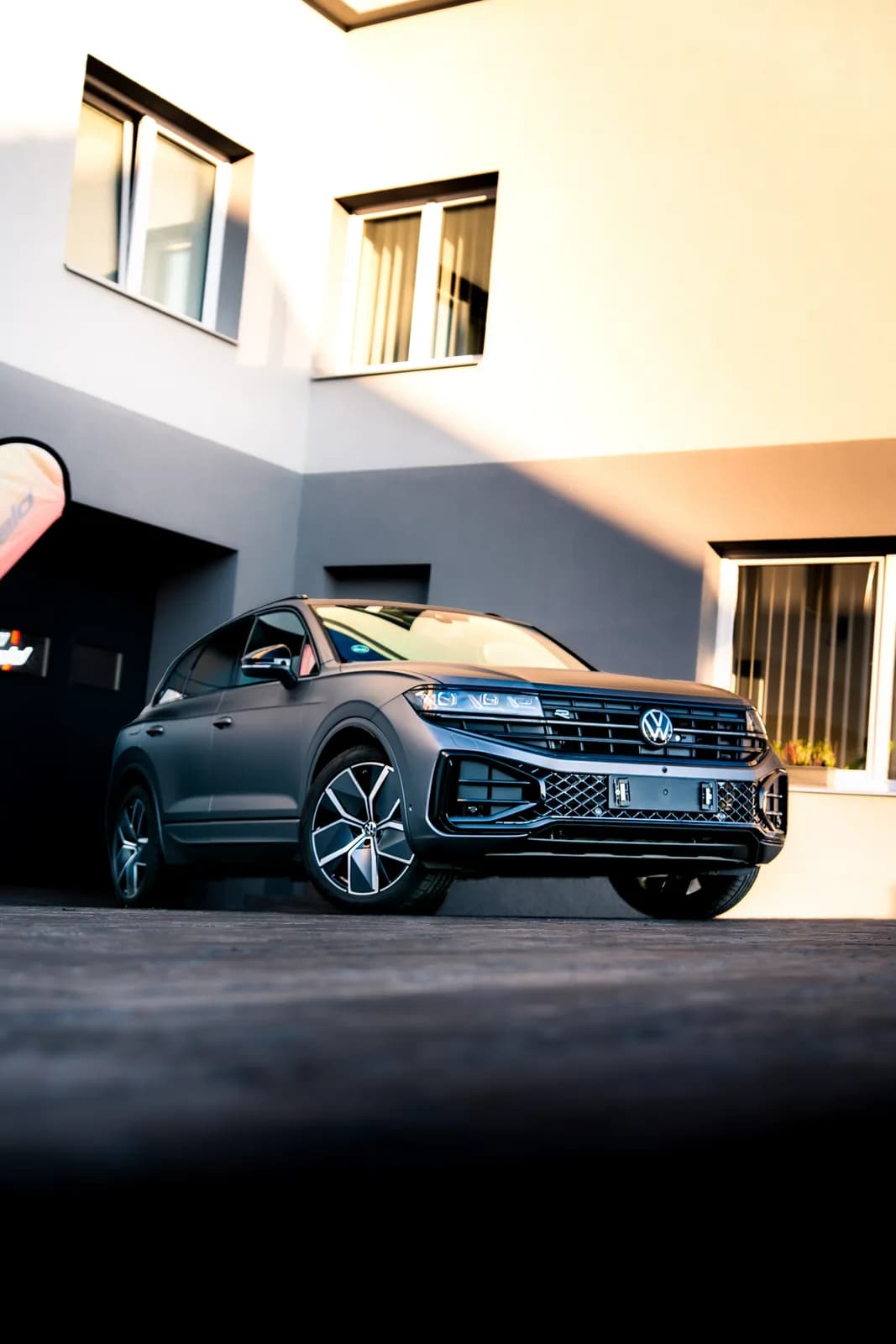 Volkswagen Touareg v matné šedé fólii zešikma zepředu z nízkého úhlu, před budovou provozovny v teplém světle.
