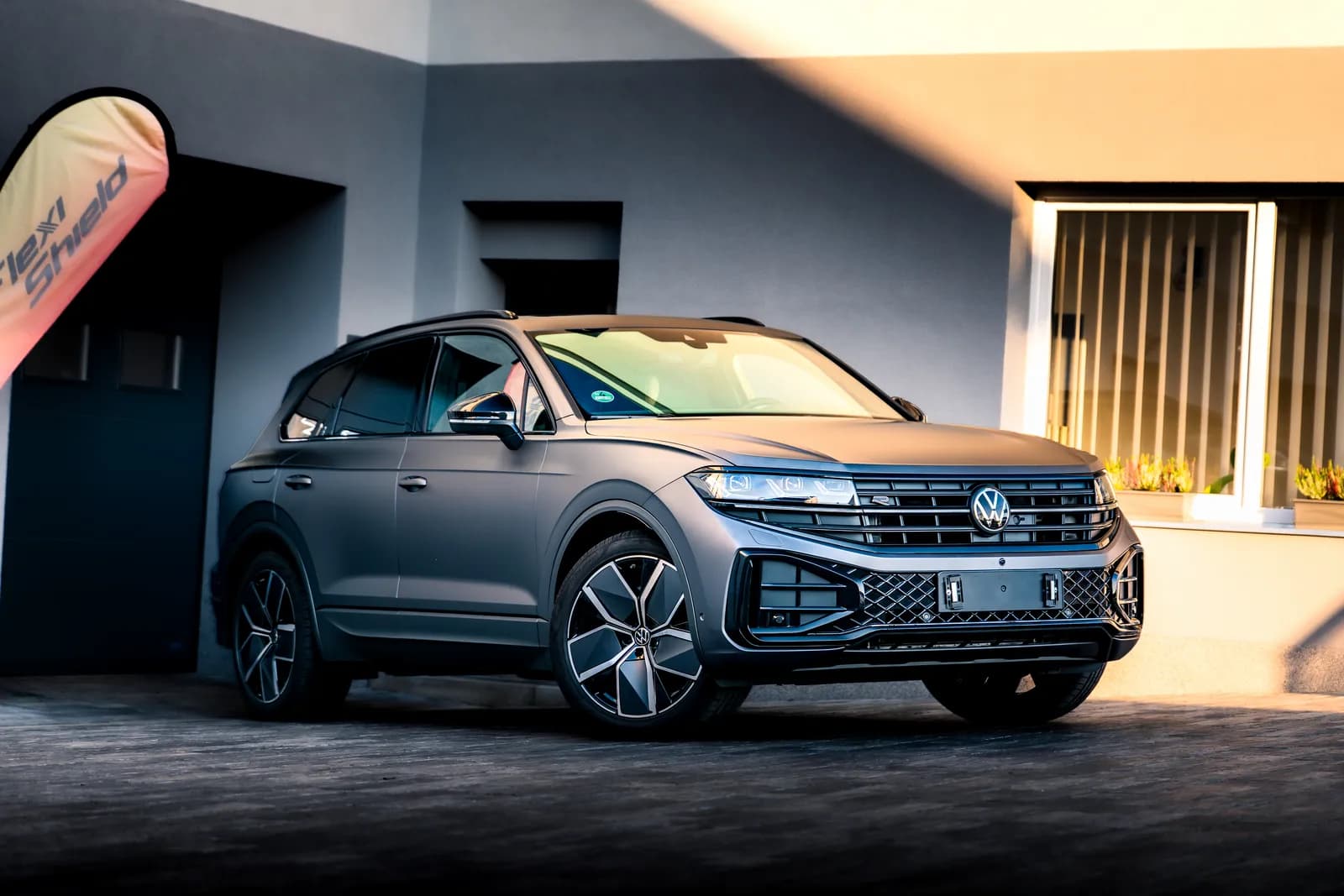Celý Volkswagen Touareg v matné šedé fólii zešikma zepředu před budovou provozovny, vpravo okno s květinami.