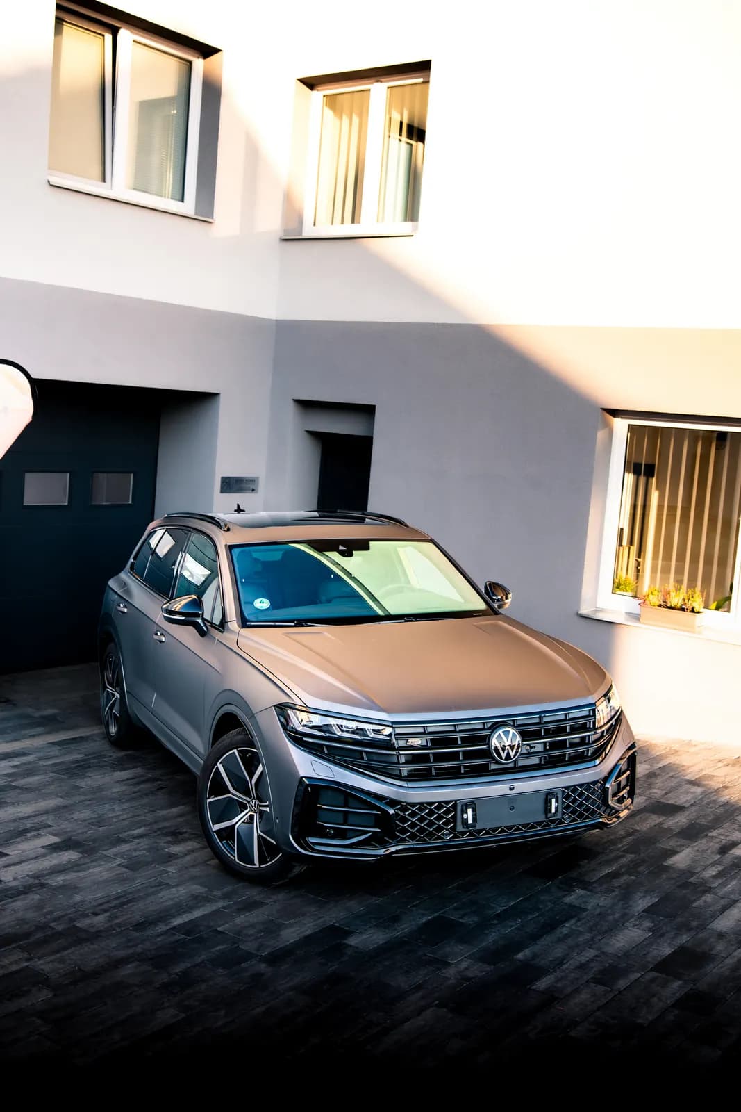 Volkswagen Touareg v matné šedé fólii zešikma shora zepředu na dlážděném dvoře před budovou.
