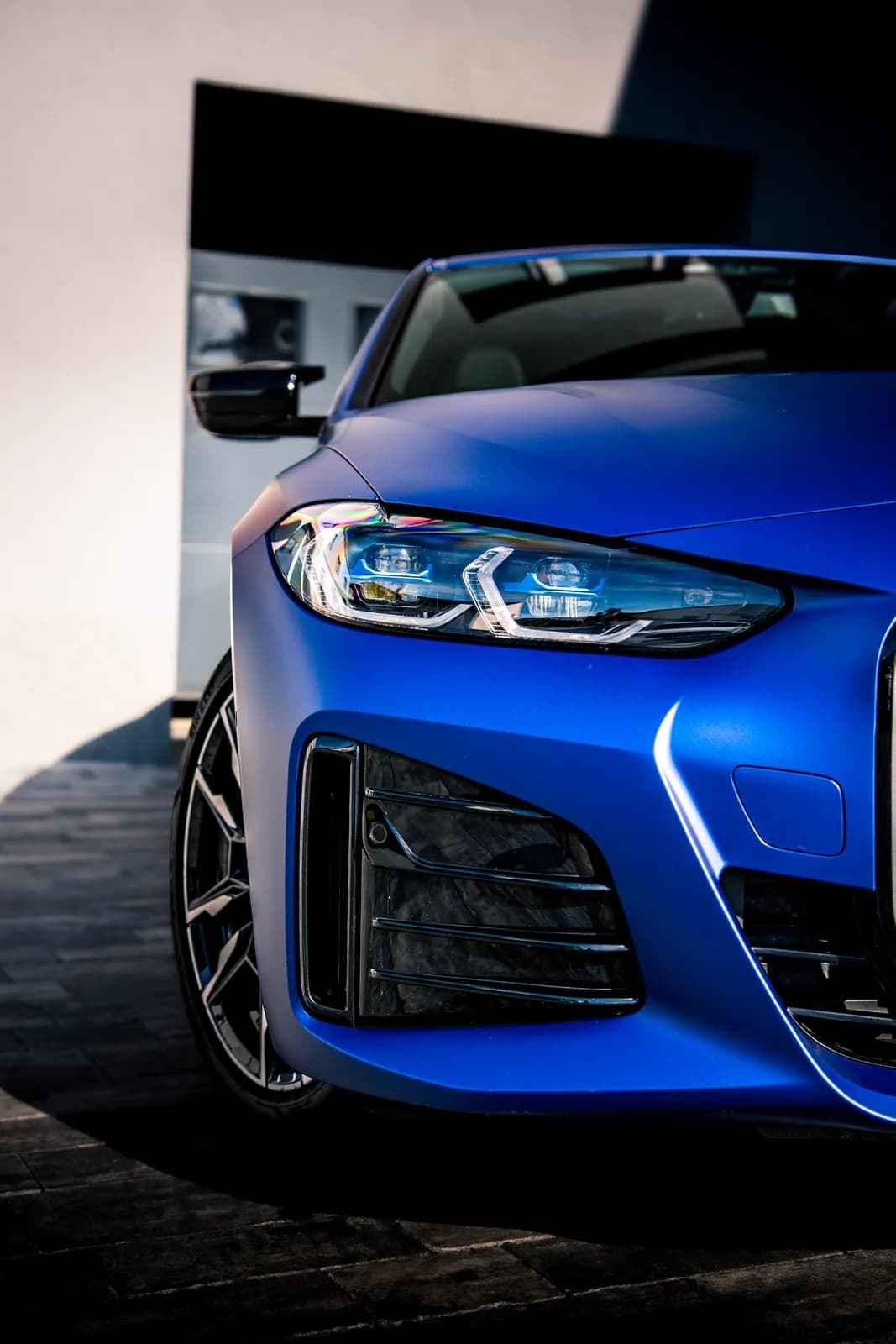 Přední část matně modrého BMW i4 zblízka: světlomet, vzduchový otvor v nárazníku a kolo.
