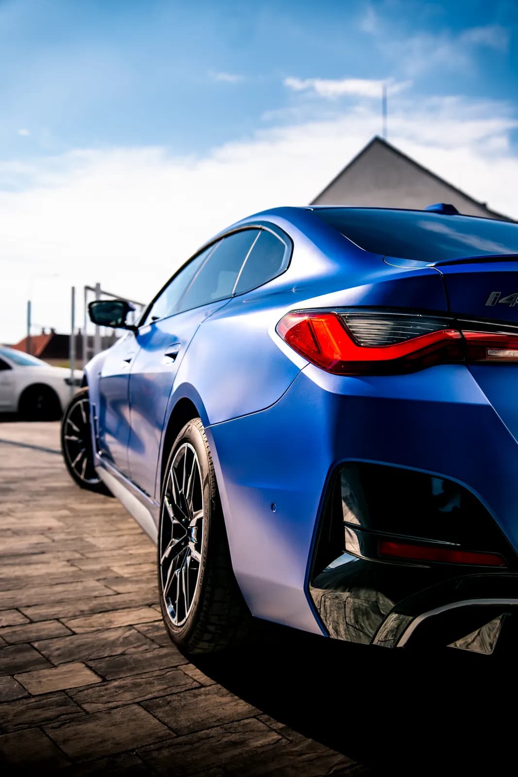 Matně modré BMW i4 zezadu a ze strany se zadním světlem, bokem a kolem; v pozadí obloha a střechy domů.
