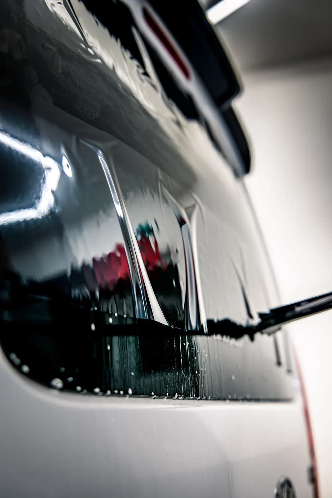 Close-up of a rear window while tint film is being applied: wet film on the glass with water droplets, the wiper and white bodywork.