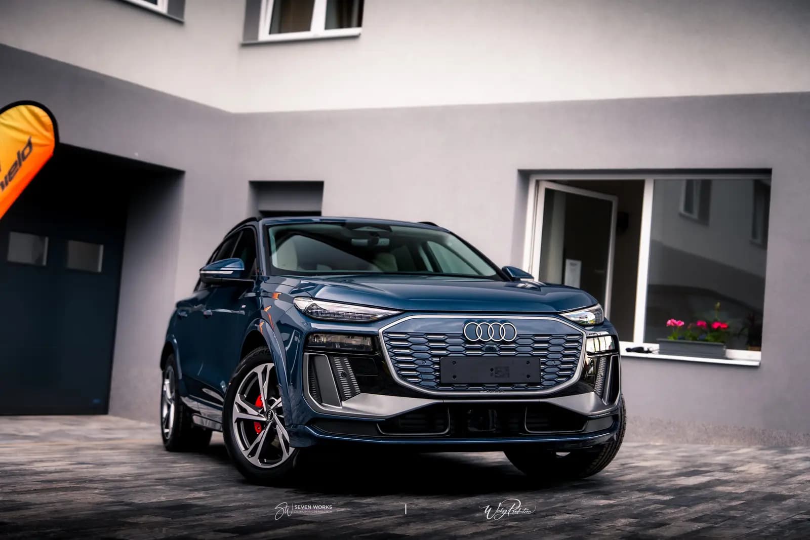 Audi Q6 e-tron zepředu u provozovny, v pozadí oranžová vlajka.