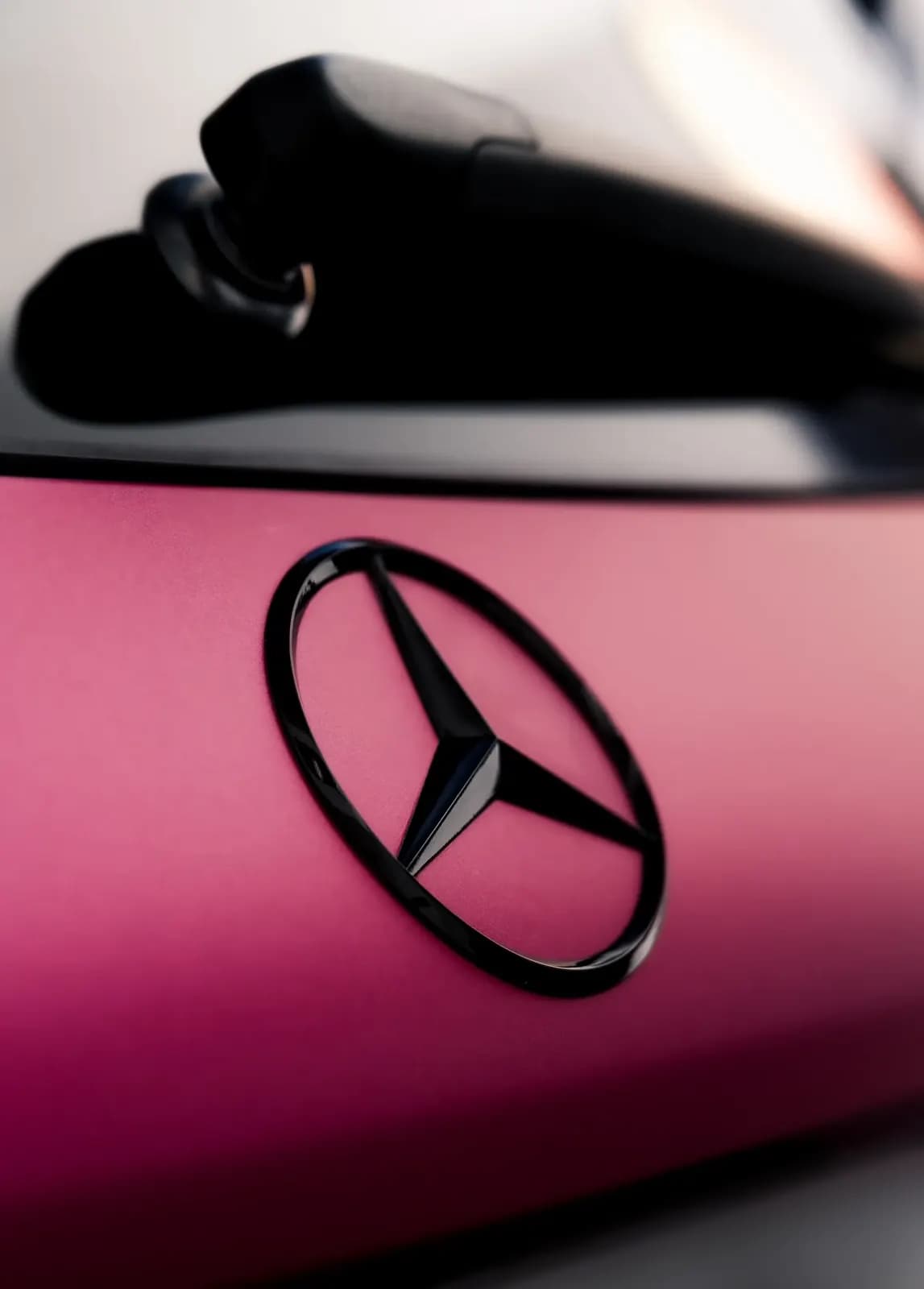 Makro detail loga Mercedes na matném růžovém polepu karoserie.