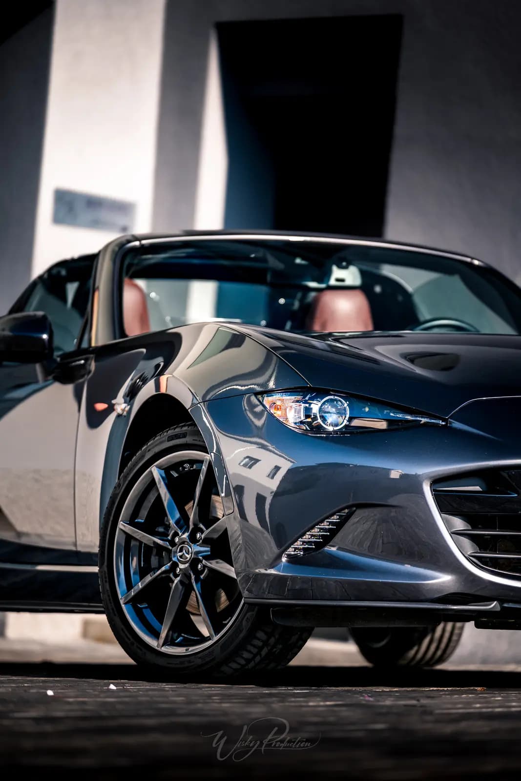 The front of a dark grey Mazda MX-5 from a low angle, with the polished wheel and headlight in frame.