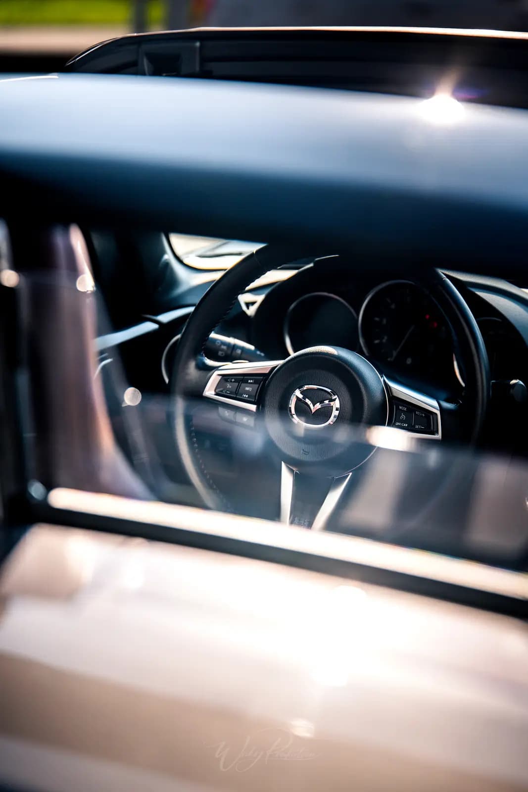 The Mazda MX-5 interior through the window: badged steering wheel, round dials and tan leather.