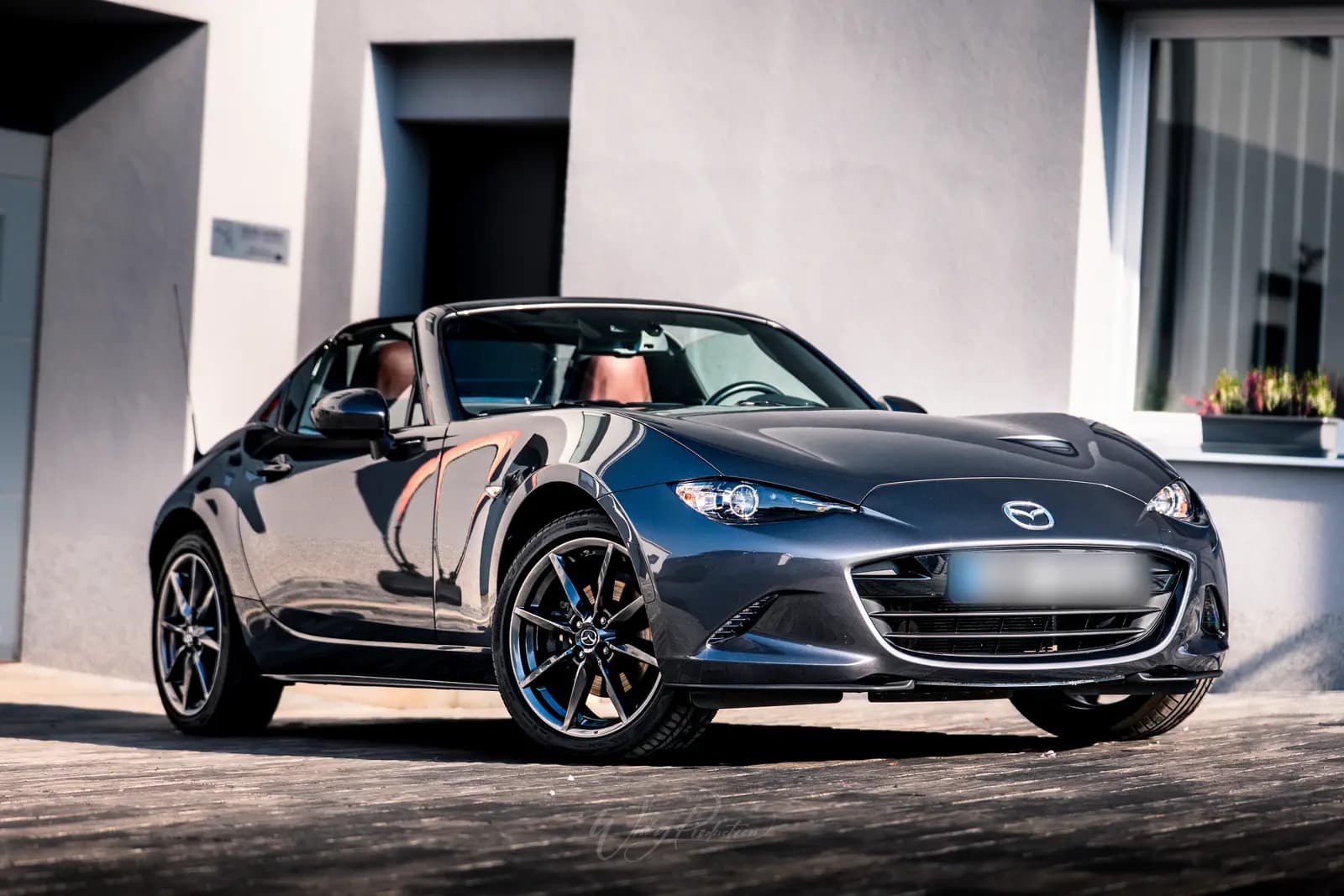 A dark grey Mazda MX-5 RF from the front quarter outside the studio, a window box of flowers behind.