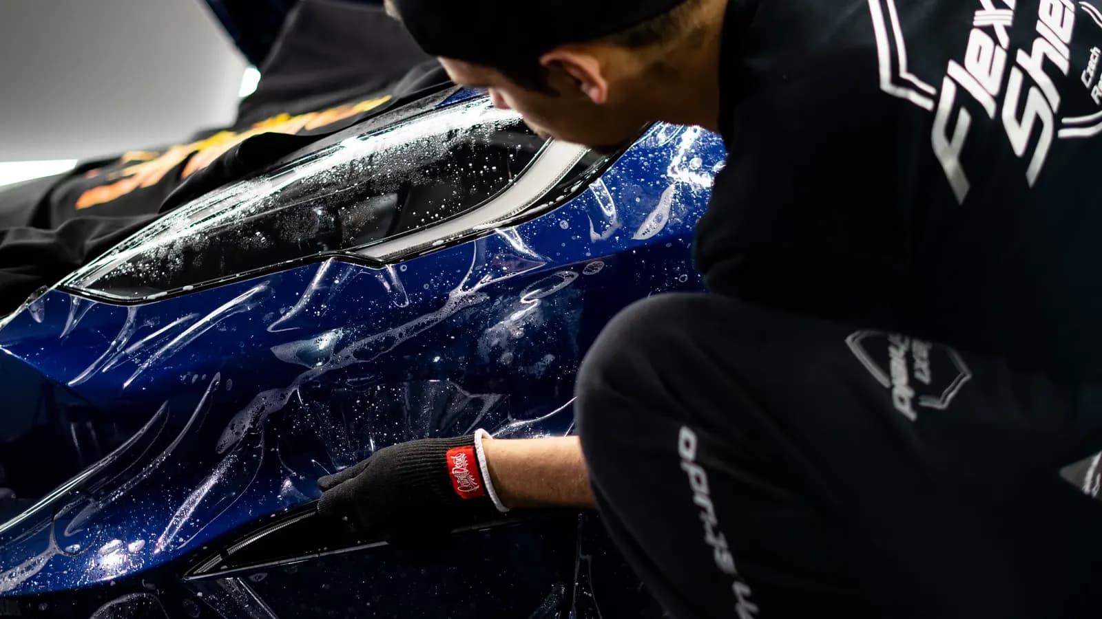 Technik v pracovním oblečení FlexiShield nanáší ochrannou fólii na přední nárazník a světlomet modrého vozu.