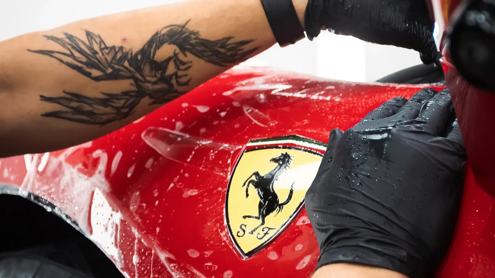 Ruce technika v rukavicích uhlazují mokrou ochrannou fólii u štítu Ferrari na předním blatníku.