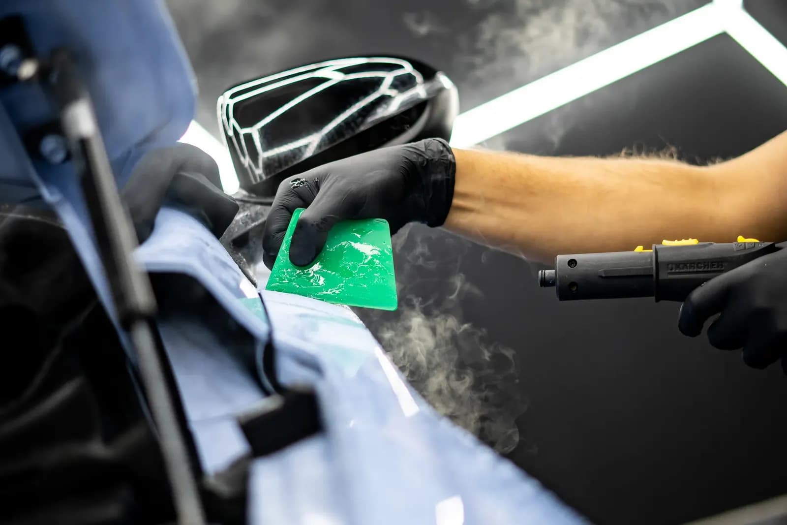 Technik zelenou stěrkou a napařovačem instaluje ochrannou fólii na světle modrý vůz.
