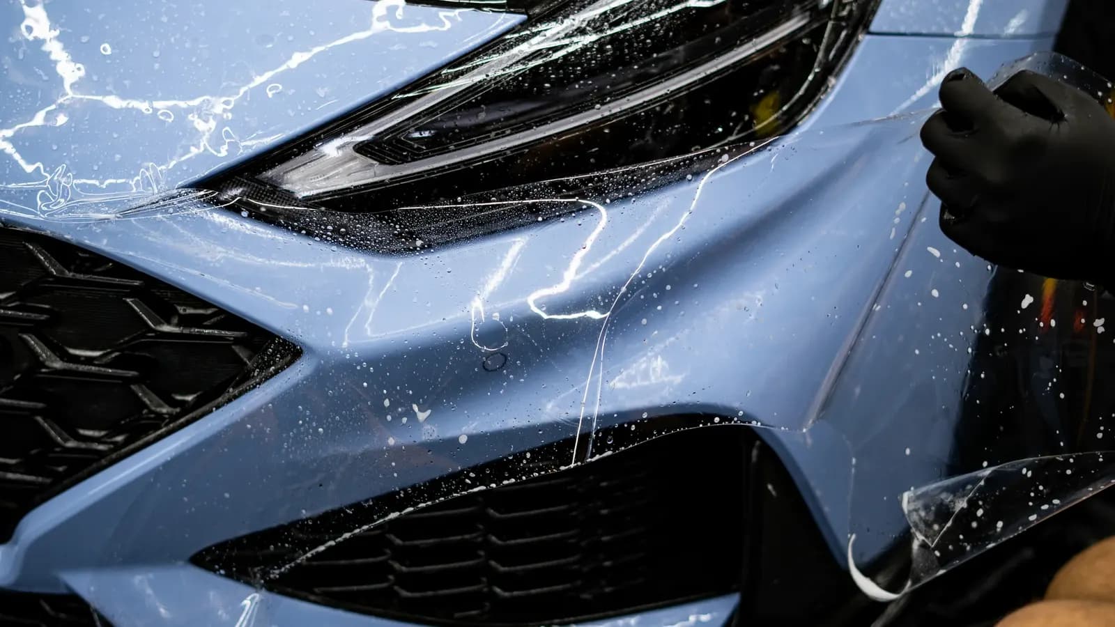 Detailní záběr ruky technika uhlazujícího mokrou ochrannou fólii na přední masce modrého vozu.