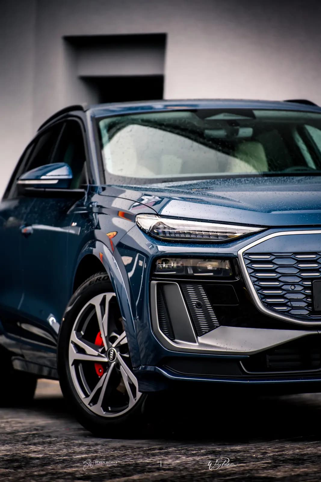 Detail přední masky, světlometu a předního kola modrého Audi Q6 e-tron u budovy provozovny.