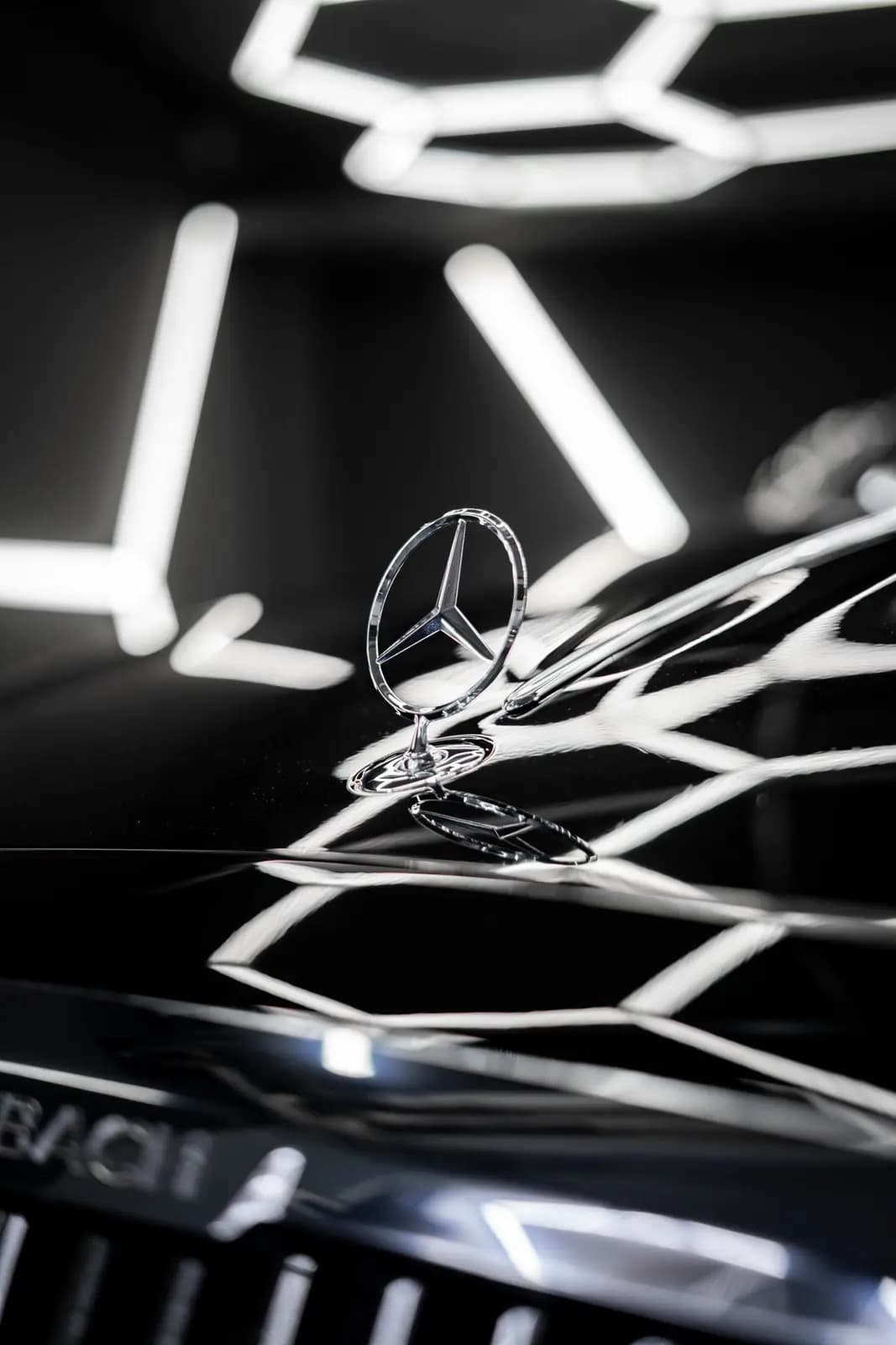 Hvězda Mercedes-Benz na lesklé černé kapotě s odrazem šestiúhelníkových světel.