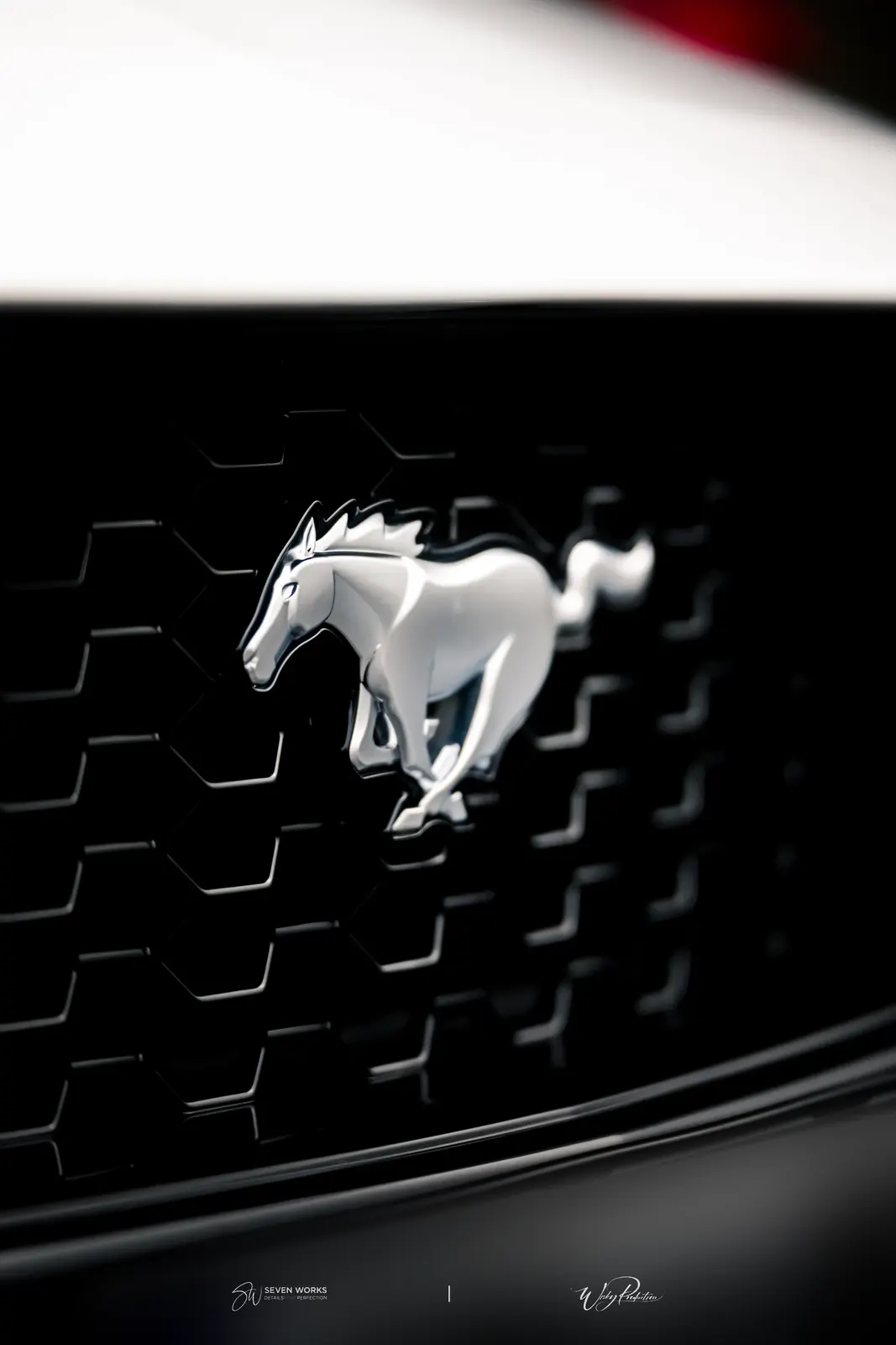 Makro detail chromovaného loga Ford Mustang na černé masce chladiče.