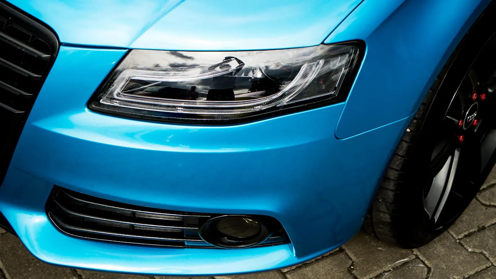 Detail předního světlometu a nárazníku modrého vozu Audi s černým kolem.