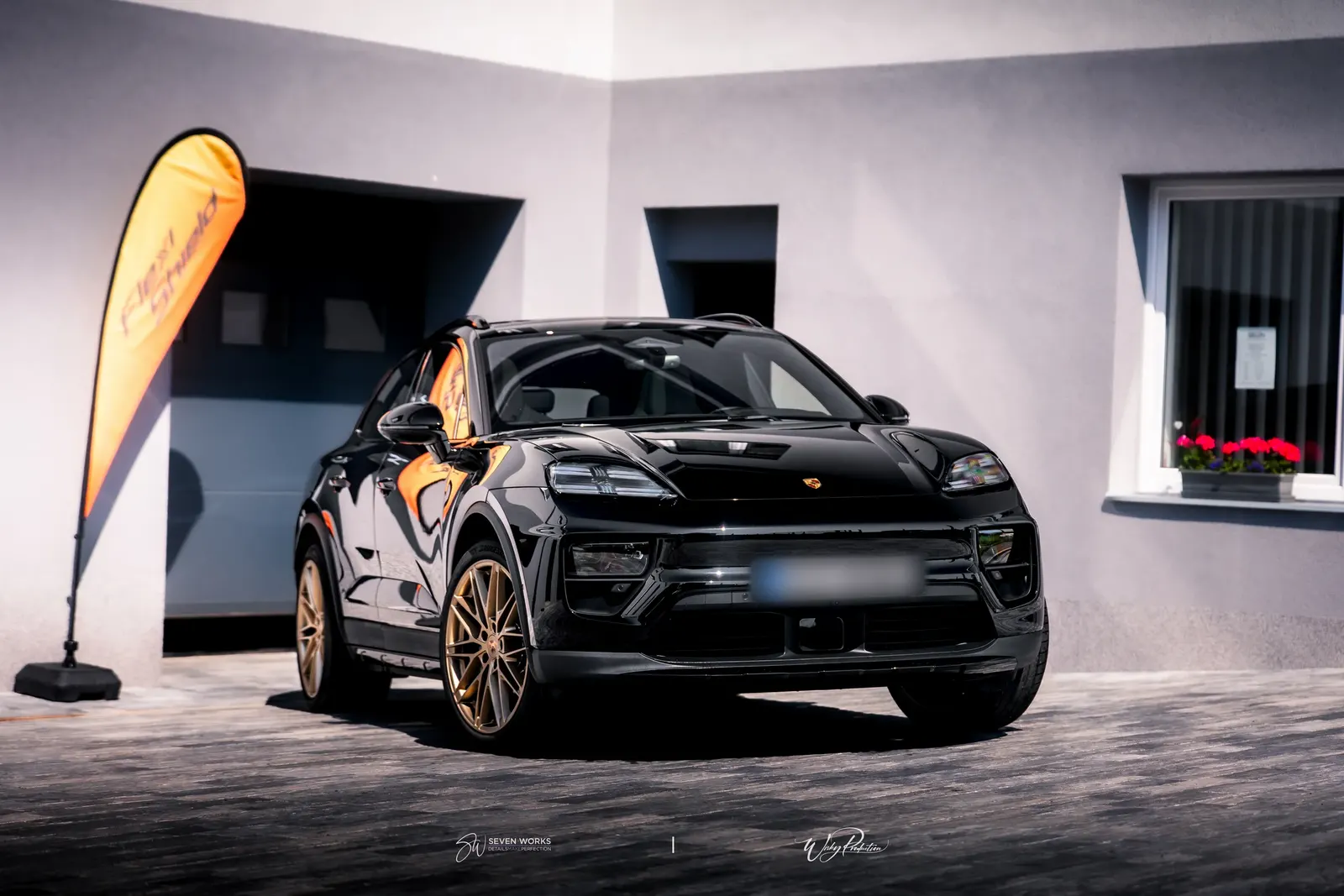 Černé Porsche s bronzovými koly před garáží, vlevo oranžová vlajka a vpravo okno s květinami.