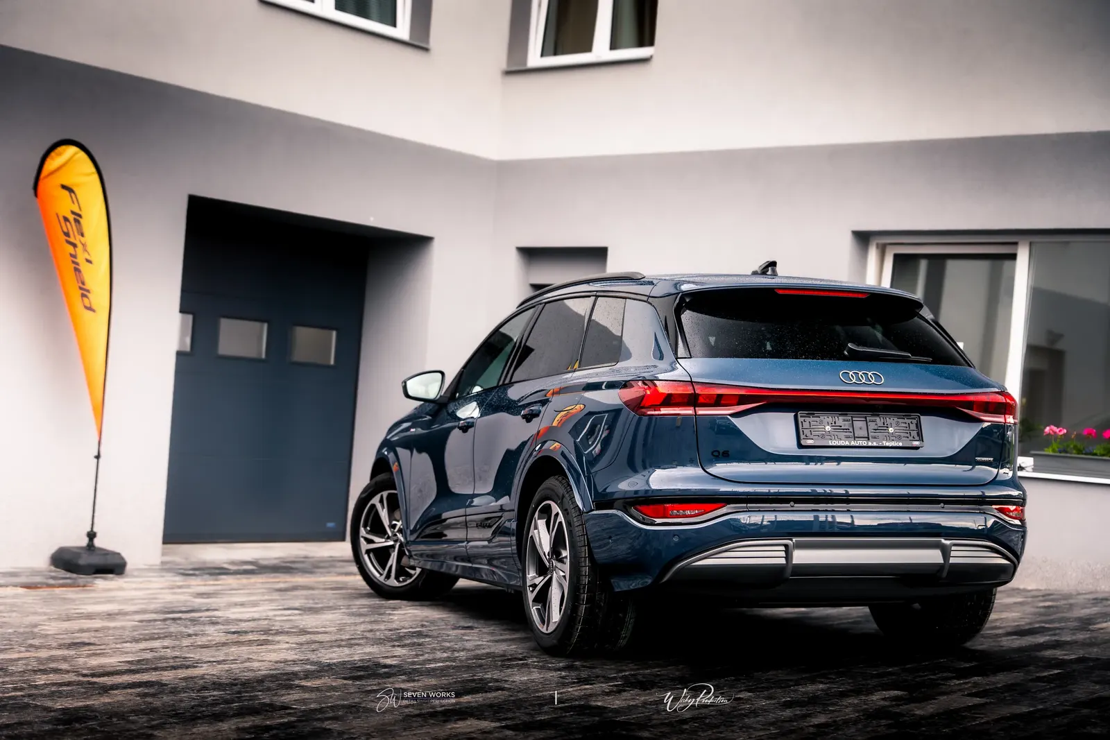 Zadní 3/4 pohled na tmavě modré Audi SUV před domem, vlevo reklamní vlajka s nápisem FlexiShield.