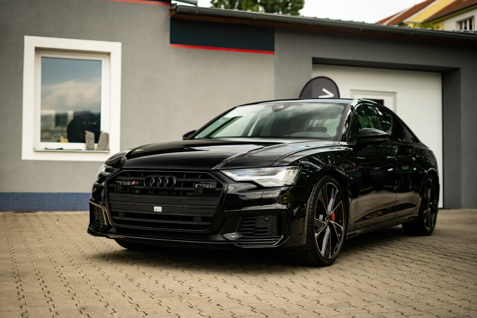 Černé Audi sedan zepředu zboku s logem Audi na masce a červenými brzdovými třmeny, zaparkované na dlažbě před šedou budovou s oknem.
