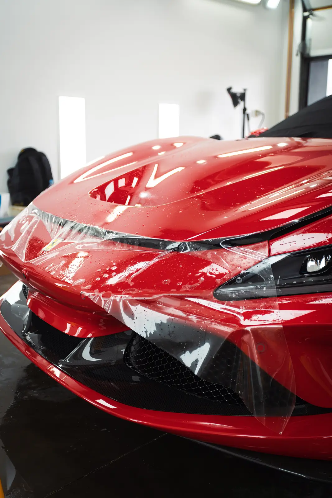 Přední část červeného Ferrari s nalepovanou ochrannou fólií na světlometu a nárazníku, v pozadí světelné panely v místnosti.