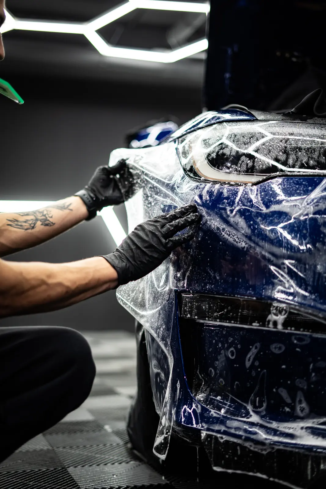 Protective film applied to the front bumper and headlight of a dark blue car, honeycomb studio lighting behind.
