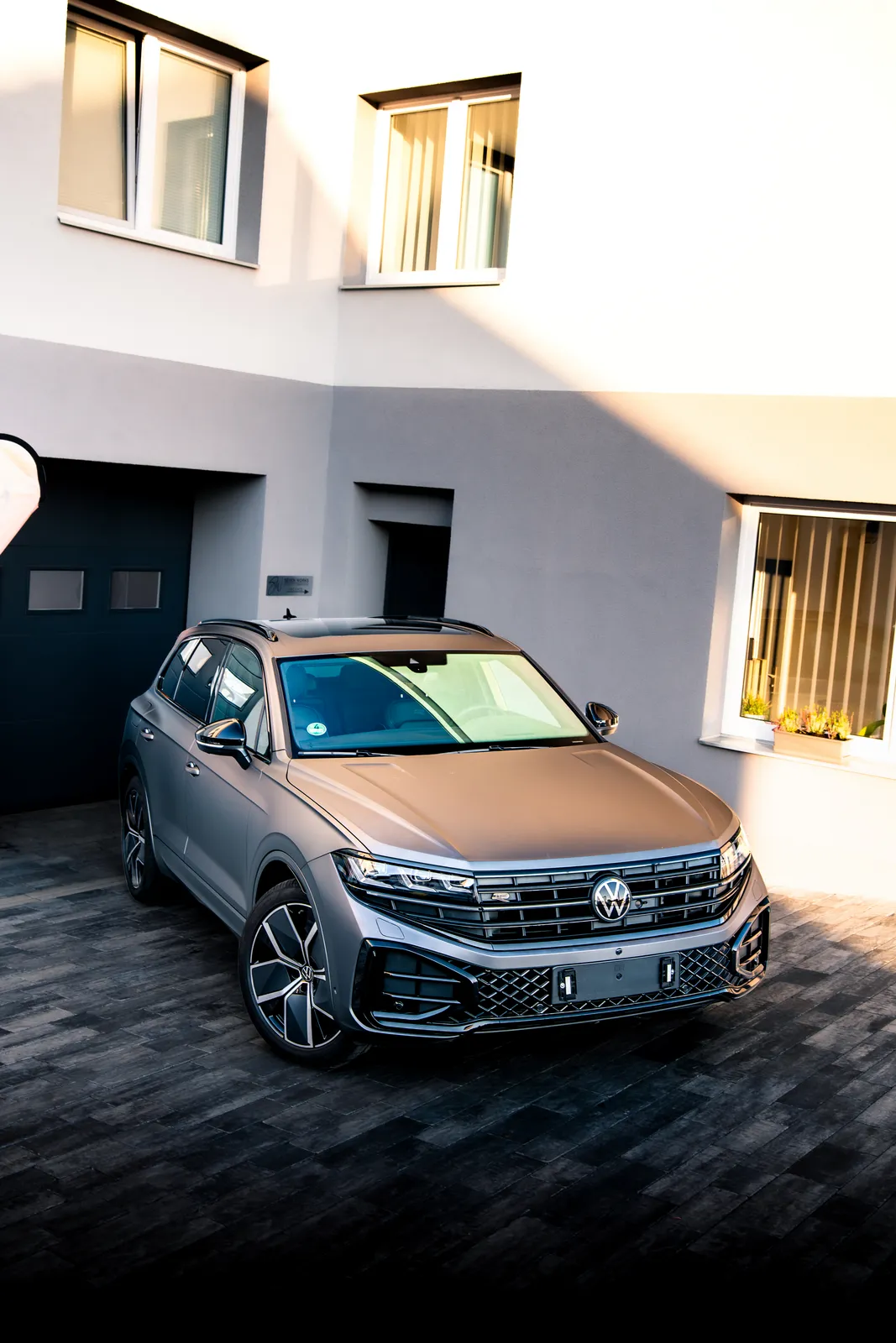 Volkswagen Touareg v matné šedé fólii zešikma shora zepředu na dlážděném dvoře před budovou.