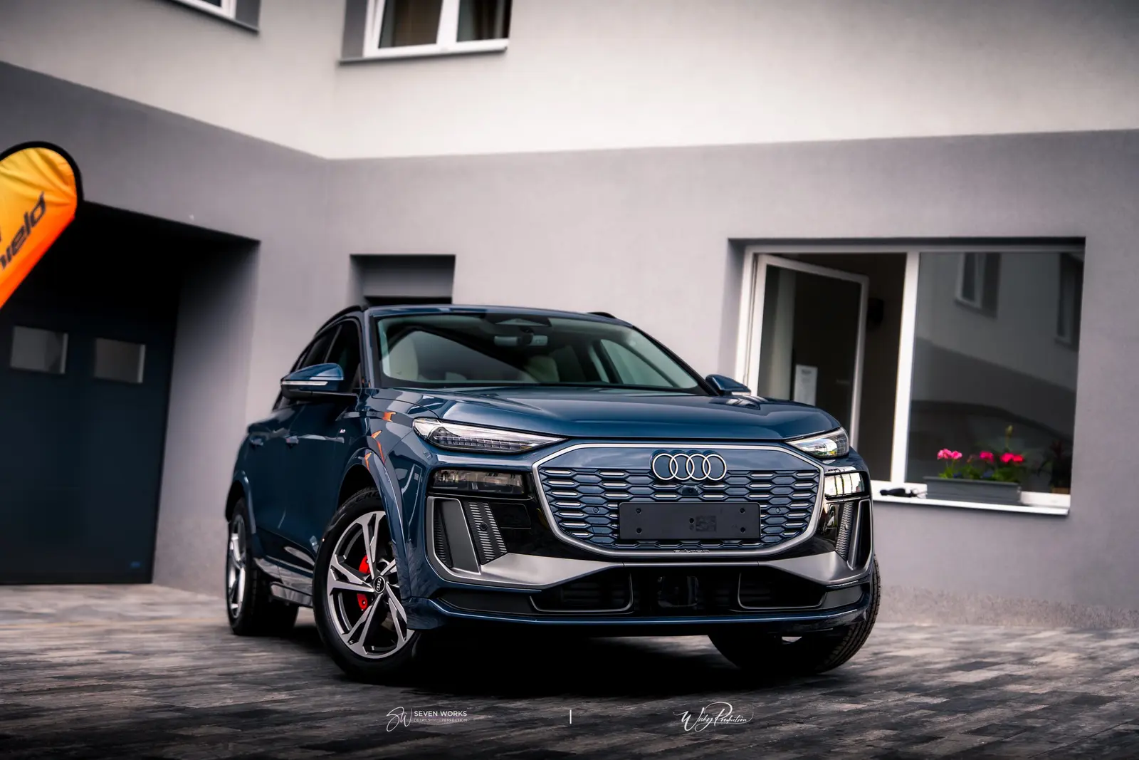 Audi Q6 e-tron zepředu u provozovny, v pozadí oranžová vlajka.