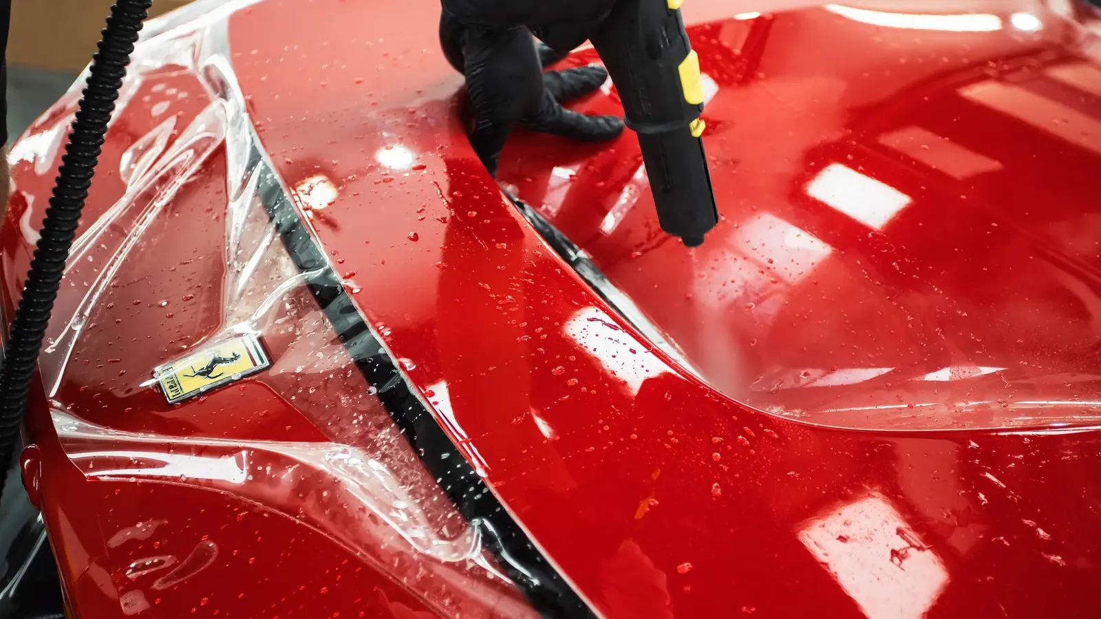 Technik v rukavicích prohřívá párou ochrannou fólii na kapotě červeného Ferrari.