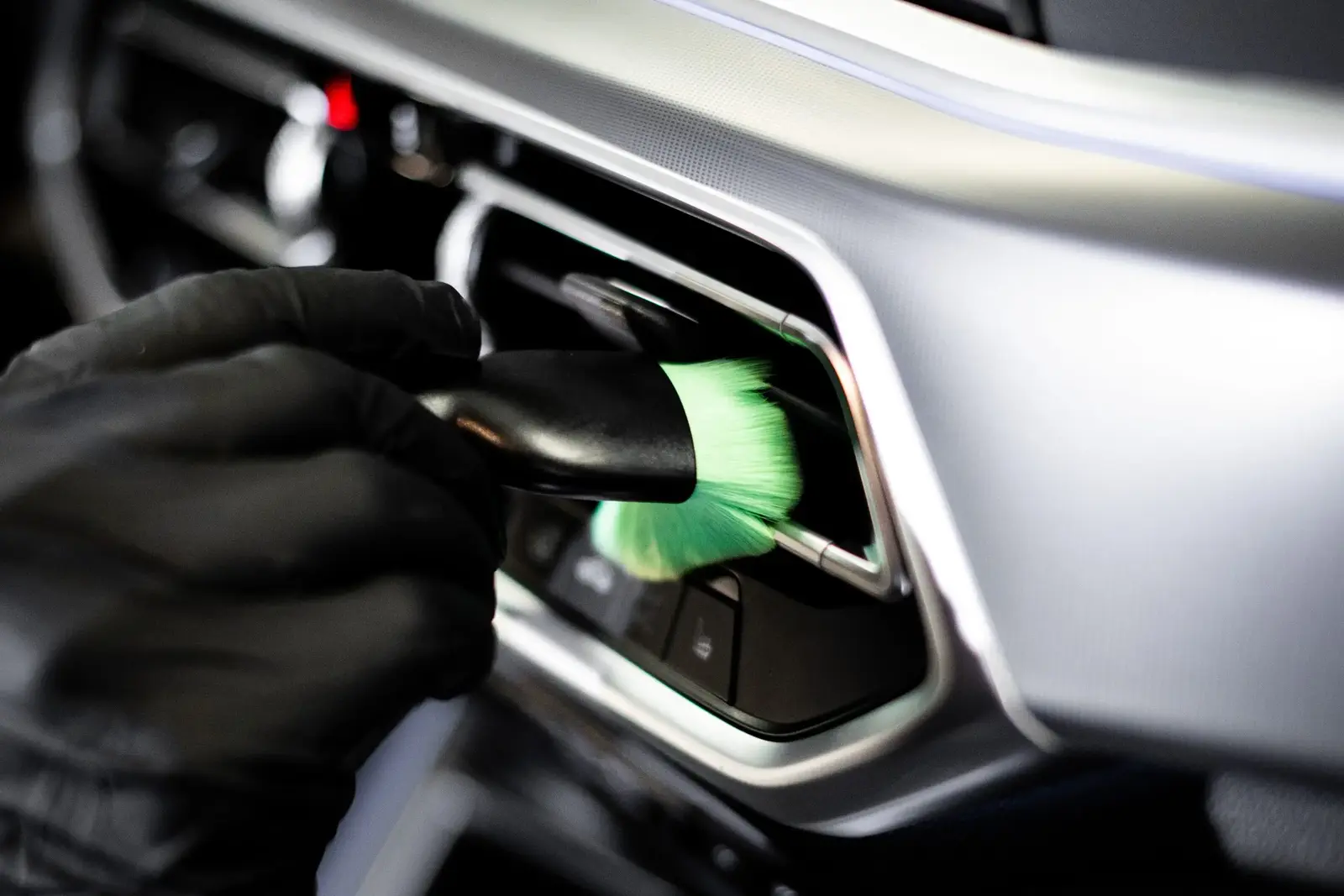 A gloved hand cleans a dashboard air vent with a detailing brush.
