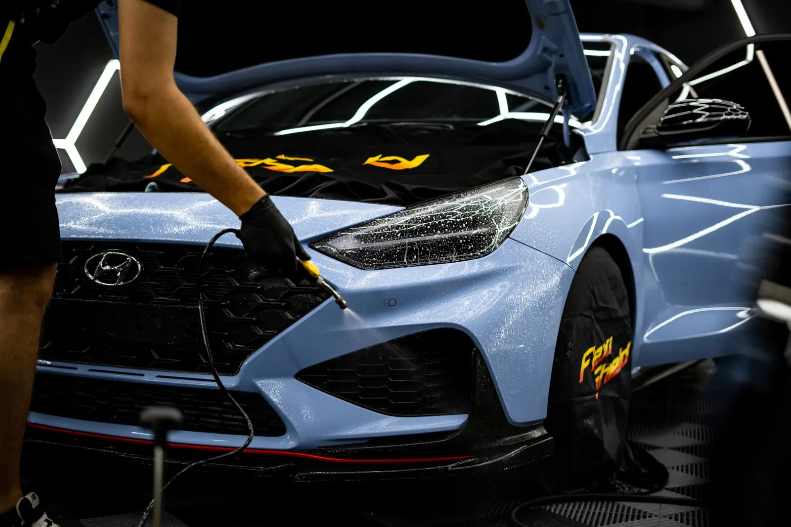 Technik stříká kapalinu z ruční pistole na přední nárazník světle modrého vozu s otevřenou kapotou.