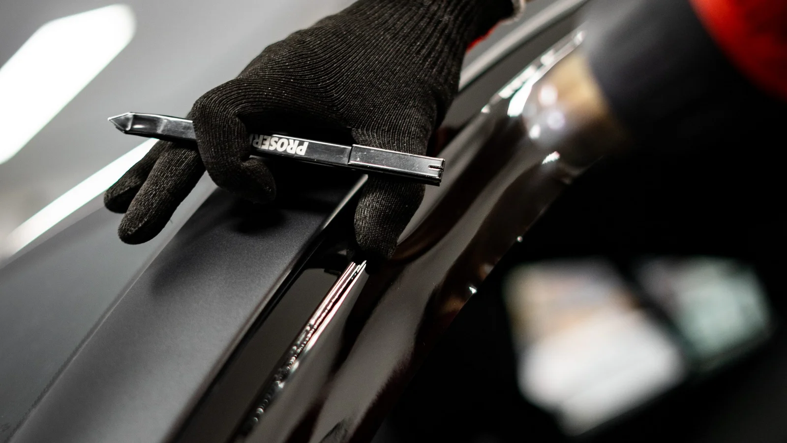 A technician's hand in a knitted glove holds a tool at the edge of matte protective film on a car body.